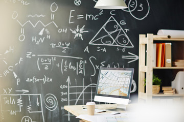Background shot of empty workplace in sunlight: desk with computer and supplies standing against blackboard with scientific formulas written in chalk