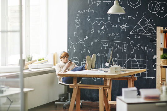Red haired man wearing beanie hat and glasses resting in chair with legs on table using smartphone against blackboard with scientific formulas written in chalk