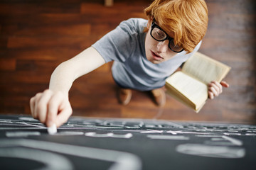 Top view portrait of young red haired man writing on blackboard with chalk holding book in hand...