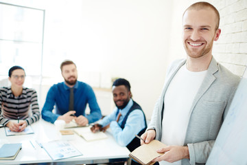 Obraz premium Portrait of cheerful young businessman in casualwear with pen and notebook in hands looking at camera while leading business meeting for his subordinates