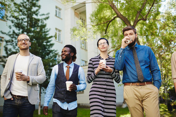 Multiethnic group of coworkers standing at office building and discussing their promising project over cup of coffee, low angle view