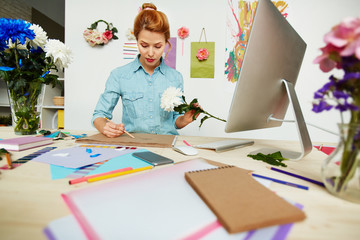 Waist-up portrait of beautiful artist in denim shirt drawing picture of white chrysanthemum with pencil, office desk with folders, notebooks, color swatch books and glass vases on foreground