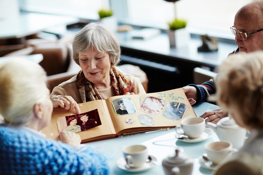 Four senior friends hanging out in lovely coffeehouse: they drinking delicious tea and listening attentively to pretty woman with worn-out photo book in hands