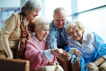 Four tech-savvy senior people gathered together and taking selfie with modern smartphone against panoramic window