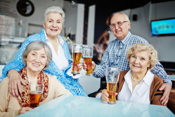 Lovely portrait of two smiling elderly women sitting in comfortable armchairs and looking at camera while their friends sitting on armrests and embracing them slightly