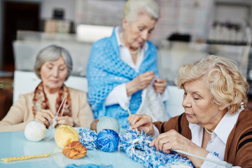 Knitting club for senior people: three concentrated women sitting at table and knitting Christmas presents for their grandchildren