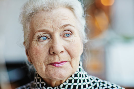 Head shot of beautiful grey-haired senior woman with deep blue eyes looking away thoughtfully, blurred background