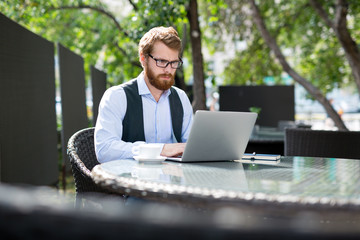 Handsome young businessman in formalwear sitting under shade of trees in small outdoor cafe and writing response letter to business partner with help of laptop