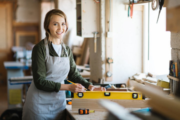 Portrait of pretty young carpenter in apron looking at camera with wide smile while holding spirit level on plank, blurred background - Powered by Adobe