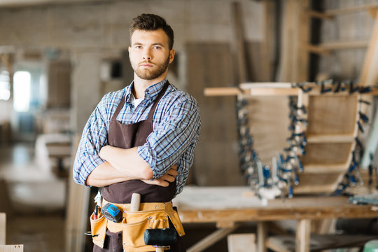 Portrait of serious young woodworker wearing leather tool belt posing for photography while standing in workshop with arms crossed