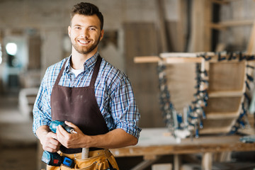 Waist-up portrait of smiling bearded craftsman with electric drill in hands standing in spacious workshop and looking at camera - obrazy, fototapety, plakaty