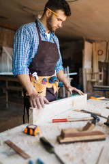 Bearded carpenter with pencil behind his ear checking surface of wooden board, table with shavings and hand tools on foreground