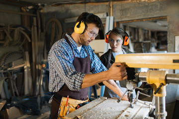 Skillful woodworker in eye and ear protectors explaining his female apprentice how to use drill press, she listening to him with attention - obrazy, fototapety, plakaty