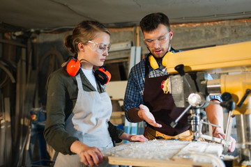 Waist-up portrait of attractive apprentice standing at drill press and listening to her male mentor...