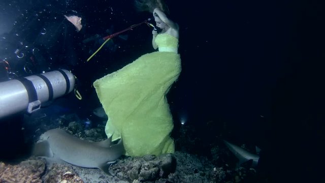 Scuba diver gives air to a young beautiful woman in a yellow dress posing underwater with a Tawny nurse sharks (Nebrius ferrugineus), a night shoot, the Indian Ocean, the Maldives
