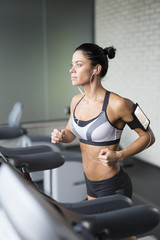 Fototapeta premium Portrait of sportive brunette woman running on treadmill in gym listening to music using shoulder smartphone holder