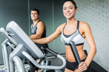 Portrait of beautiful  sportive brunette woman exercising using elliptical machine  next to fit man, both smiling to camera during workout in modern gym