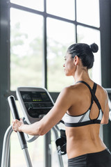 Fototapeta na wymiar Back view portrait of beautiful sportive woman exercising using elliptical machine during workout in modern gym against big window