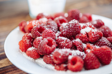 Ripe raspberry on a white plate powdered with sugar. Plate on a wooden table. Glass of milk at the background.