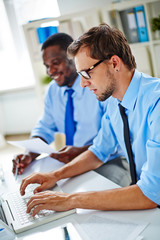 Obraz premium Profile view of handsome bearded businessman using laptop while his cheerful Afro-American colleague taking necessary notes in open plan office