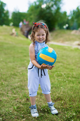 Obraz premium Pretty little girl with curly hair and headband standing on green lawn in park, holding ball and smiling at camera cheerfully