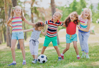 Little kids in summer wear standing in front of ball in park, embracing each other and smiling cheerfully