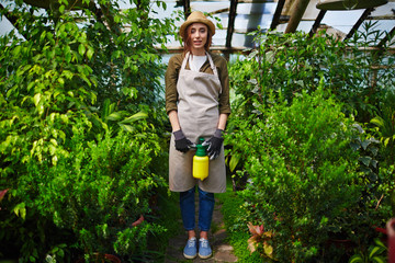 Portrait of beautiful young woman wearing straw hat and apron enjoying work in glasshouse, standing full height and looking at camera smiling between tree shrubs
