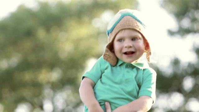 Cute happy baby of 1 year old in strong hands of father. Child wearing funny knitted pilot helmet. Daddy plays with son active games outdoors in sunset light. Real time full hd video footage.
