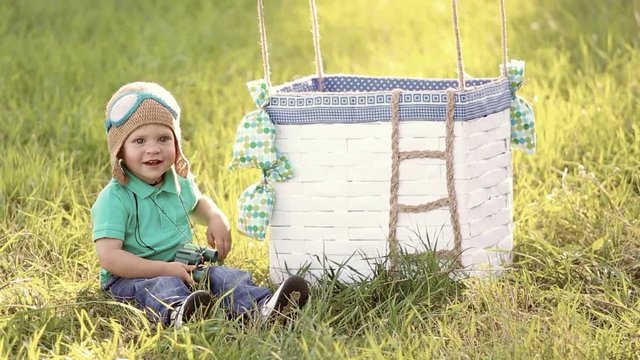 Beautiful funny little child wearing knitted pilot hat sits near handmade toy airballoon in green grass. Portrait of cute baby looking at camera smiling happily. Real time full hd video clip.