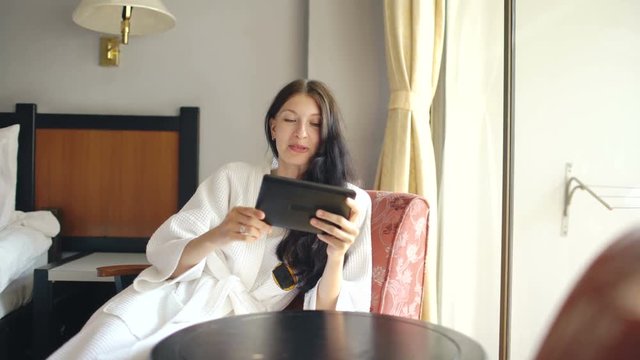 Pretty young woman in bathrobe chatting on tablet computer sitting on chair in hotel room