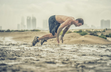A powerful strong athletic male does a flying pushup in a muddy puddle with a rough terrain with a city in the background