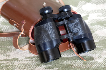 Vintage Porro prism black color military binoculars and brown leather carry case with strap on camouflage background side view close up - Powered by Adobe