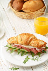 Fresh sandwich-croissant with ham, arugula, cucumber and cheese in a white plate, lemonade, orange juice and coffee Cup on the table. Breakfast. Selective focus