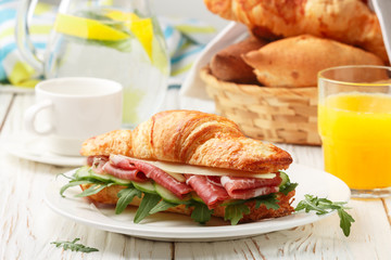 Fresh sandwich-croissant with ham, arugula, cucumber and cheese in a white plate, lemonade, orange juice and coffee Cup on the table. Breakfast. Selective focus