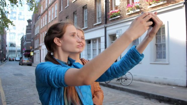 Two happy young girls fooling around taking selfie and talking . Walking the streets of London, laughing and joking. On summer vacation.