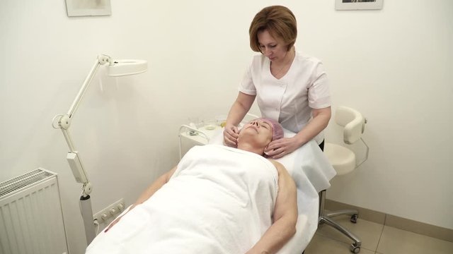 Beautician is making a face lifting procedure for an aged woman