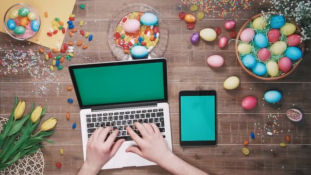 Man using laptop computer and digital tablet with green screen on table decorated with easter eggs Top view