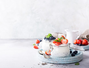 Chia Pudding for breakfast in glass jars garnished with berry in two portions on light gray stone background with a napkin and spoons. Copy space.