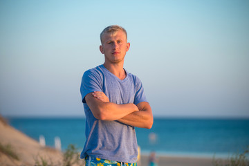 Young blond guy in a blue T-shirt and shorts stands on the background of the sea