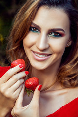 Young beautiful happy funny girl with red dress and makeup holding strawberry in summertime in the park. healthy lifestyle, diet beauty and happiness concept.