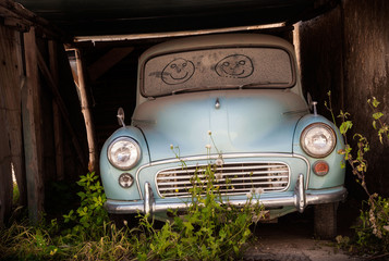 Abandoned classic car in overgrown garage with smiling faces drawn in the dusty windscreen