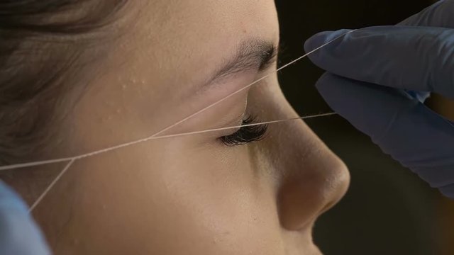 Stylist does beautiful eyebrows to the young beautiful woman. Beauty shop. Plucking out of hair thread. Close up.