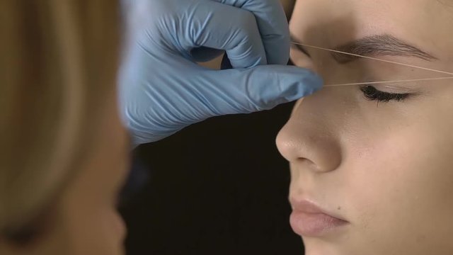 Stylist does beautiful eyebrows to the young beautiful woman. Beauty shop. Plucking out of hair thread. Close up.