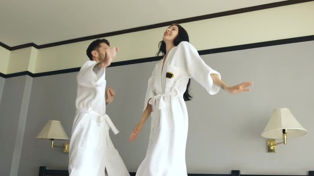 Slow motion of young happy couple in bathrobe jump and dance on bed in hotel during their honeymoon vacation