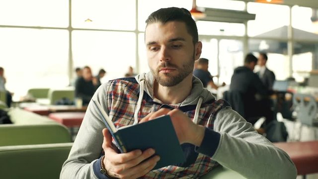 Handsome man in hoodie reading book in the cafe and smiling to the camera, steadycam shot
