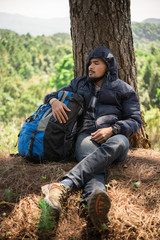 Portrait of young beard man sitting alone by a tree with backpack looking away enjoy with nature.