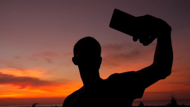 Young Man Making Selfie at Beautiful Sunset. Amazing Person Silhuette Using Mobile Phone. HD Slowmotion. Thailand.