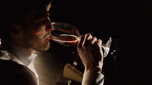 The side portrait of the man digustating the red wine at the dark blurred background. Close-up.