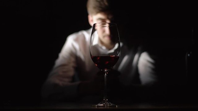 The depressed man is sitting behind the glass full of the red wine in the dack room.
