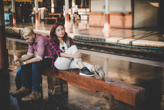 Young hipster couple sitting on wooden bench at train station. Couple sitting waiting for the train at platform.